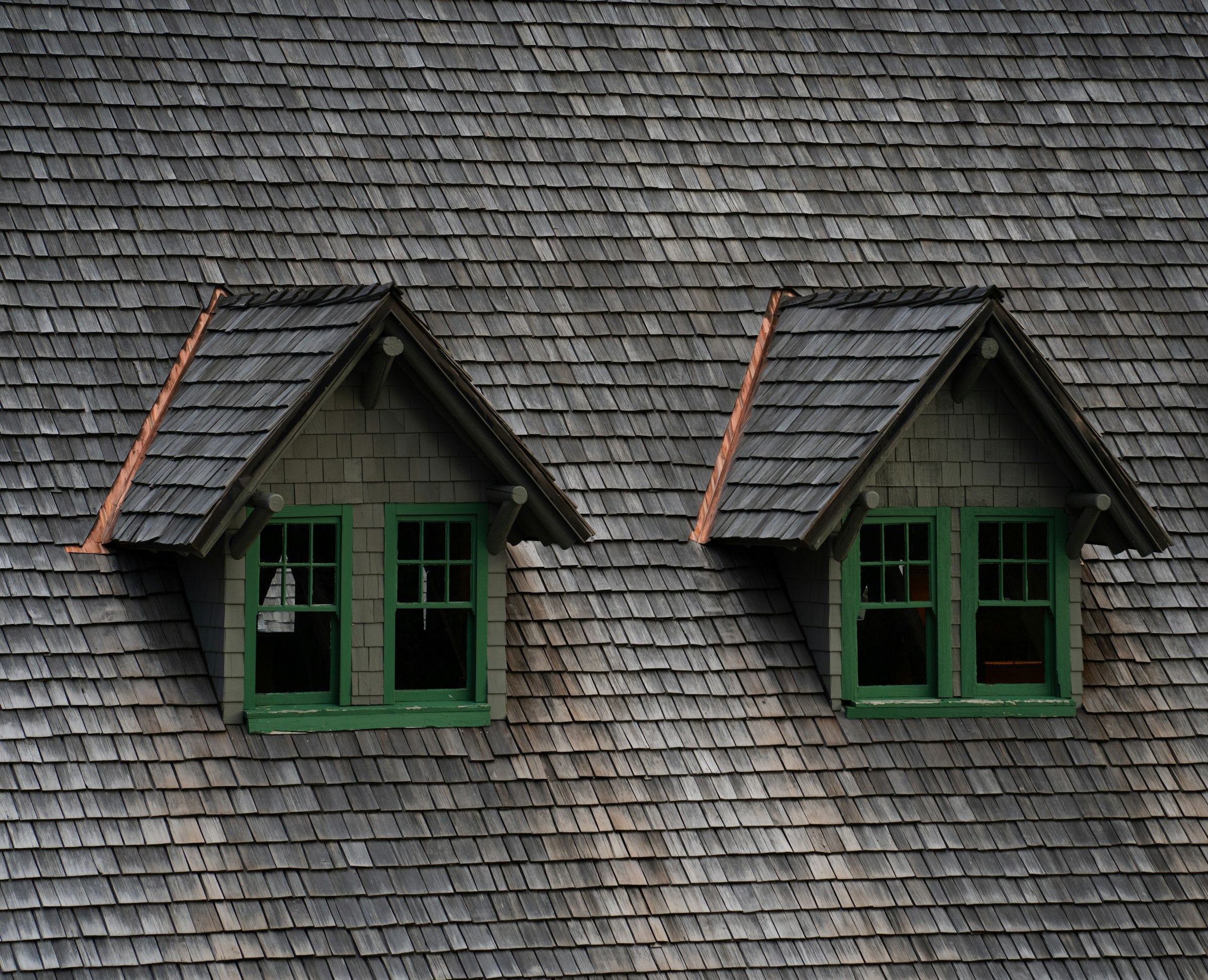 Close view of a weathered grey wood-shake roof with two shake-covered dormers, green-framed windows and copper flashing