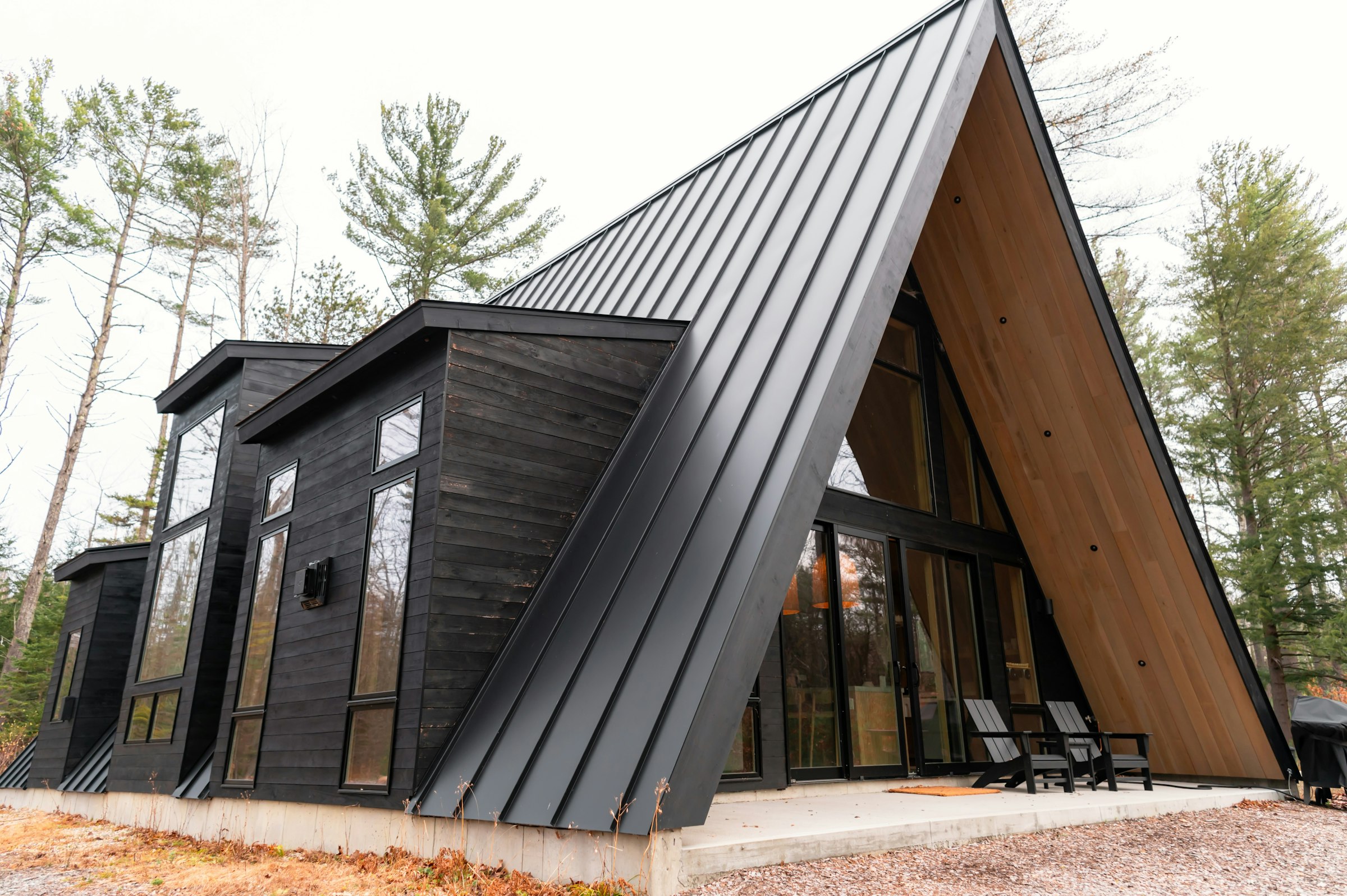 A black A-frame house whose steep dark standing-seam metal roof sweeps down beside a wood-lined glass gable, with tall pines