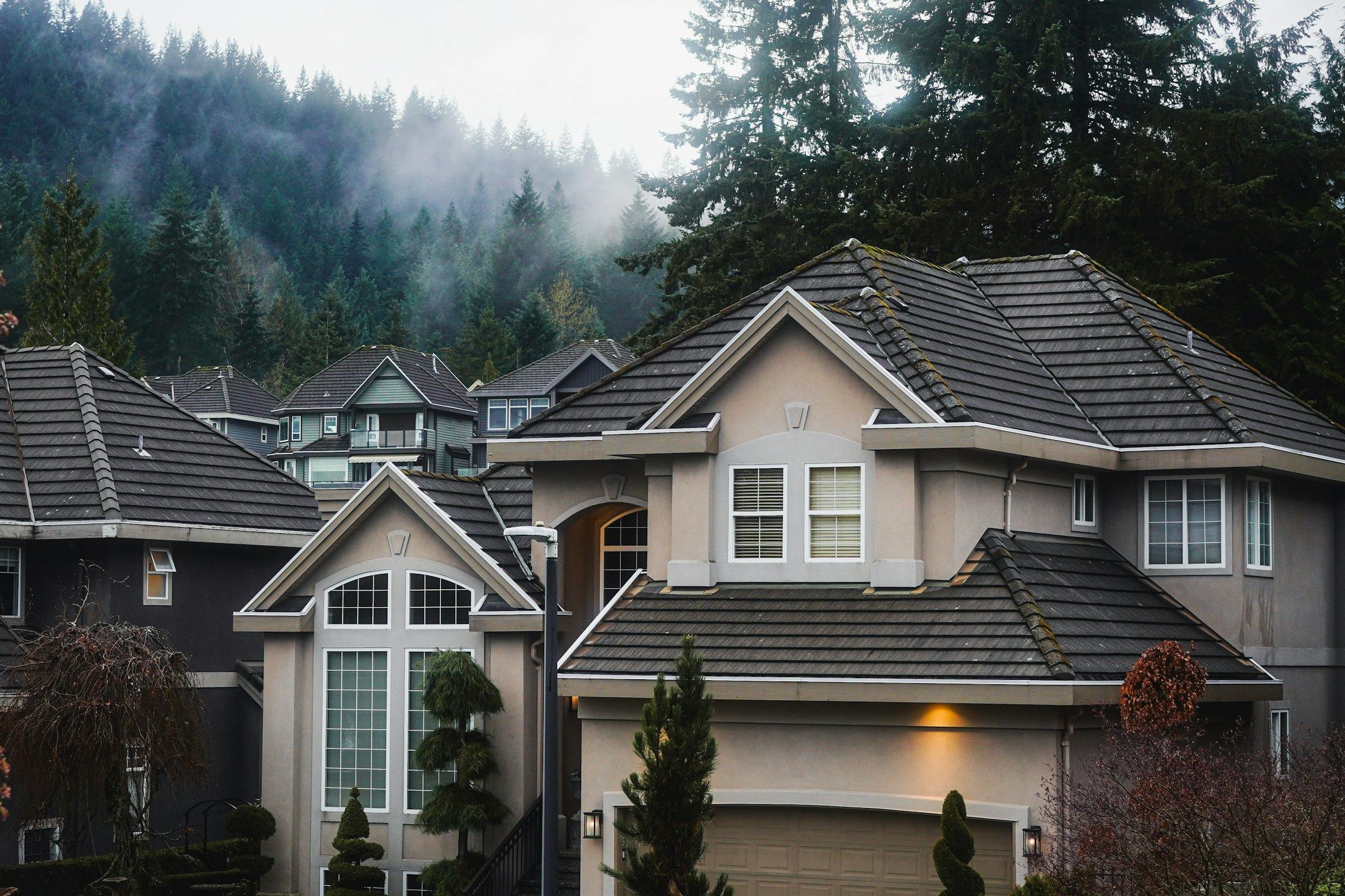 Stucco houses with dark grey flat tile roofs and white trim in front of a misty hillside of evergreen trees
